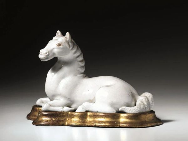  Cavallo Cina sec. XX , in porcellana bianca, in posizione accucciata, e poggiante su base in legno dorato, lung. cm 23