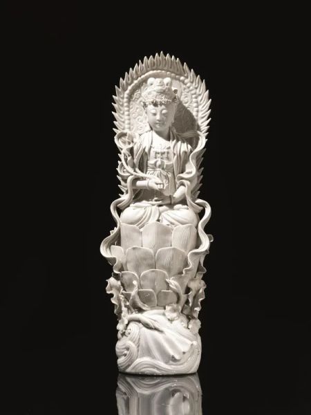 Guanyin Cina sec. XX, in porcellana blanc de Chine, assisa su fior di loto e reggente fior di loto, alt. cm 41