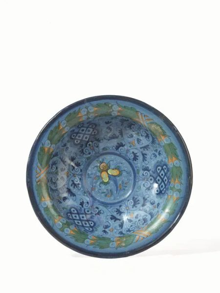 Ciotola, Faenza, met&agrave; del sec. XV, in maiolica, interamente dipinta a fondo azzurro berettino, cavetto decorato con cornucopia di frutti circondata da un fitto motivo a ramage e gruppi di losanghe blu lumeggiati da fitta puntinatura bianca e elementi minori in verde e ocra, tesa decorata da ghirlanda di frutta e foglie nei toni del verde, ocra ed azzurro, labbro sottolineato in blu, sul verso motivi decorativi in blu. Il decoro tipico delle manifatture fiorentine della prima met&agrave; del secolo &egrave; noto come motivo &ldquo;a vaghezze e gentilezze&rdquo;, diam. cm 24,5, restauri.