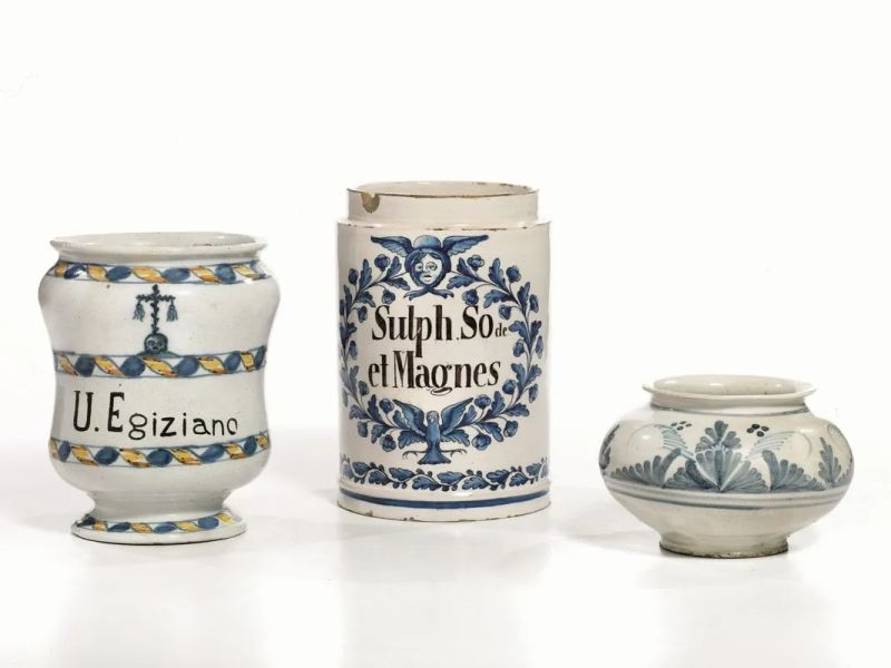 Vaso cilindrico, Italia Settentrionale o Francia, inizi sec. XIX, in maiolica, decoro con ghirlanda in blu cobalto centrata, in alto, da mascherone alato e in basso da colomba in volo; lungo il bordo inferiore una sottile ghirlanda fogliata corre lungo tutto il perimetro, alt. cm 18, privo di coperchio; albarello, sec. XIX, in maiolica, decoro a nastro continuo blu e giallo, al centro scritta farmaceutica in caratteri capitali U. EGIZIANO, alt. cm 16, restauri; pilloliere, Italia Settentrionale, sec. XIX, in maiolica, decoro a monocromia blu con fiori stilizzati, alt. cm 9 (3)  - Auction European Furniture and Work of Arts - Pandolfini Casa d'Aste